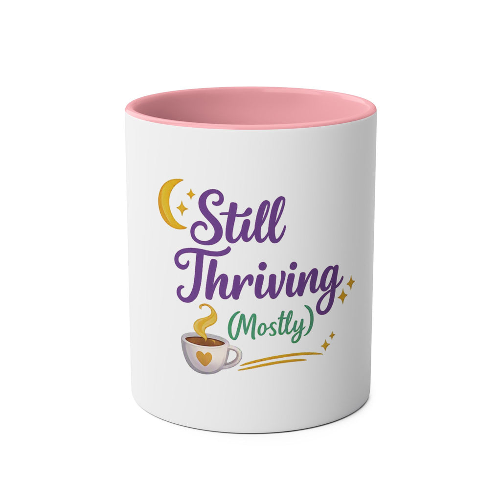 A white ceramic mug with a pink interior, featuring a graphic design on the front that reads 'Still Thriving (Mostly)' in purple and green lettering, accompanied by a yellow crescent moon, stars, and a small coffee cup illustration.