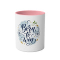 A white ceramic mug with a pink interior, featuring a graphic design on the front that reads 'Born to Win' in dark blue script over a light blue watercolor splash, decorated with gold stars, compasses, and a small green leaf.