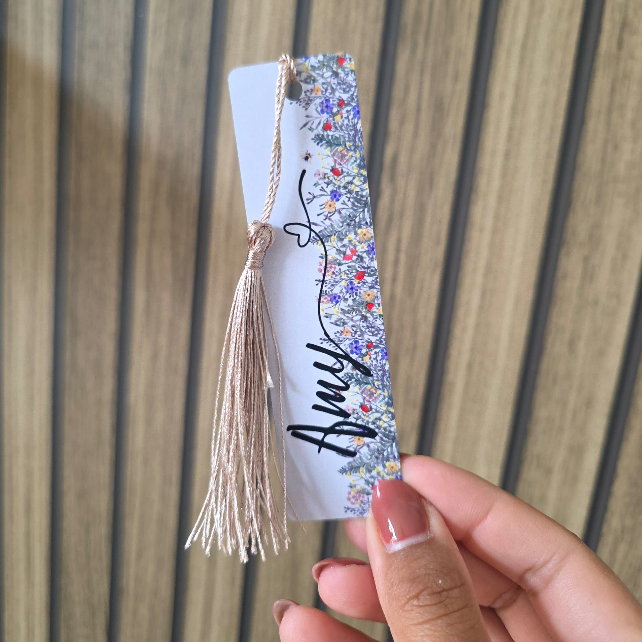 A hand holding a personalized paper bookmark with a floral patterned edge, a beige tassel, and the name 'Amy' written in black script, set against a wooden slatted background.