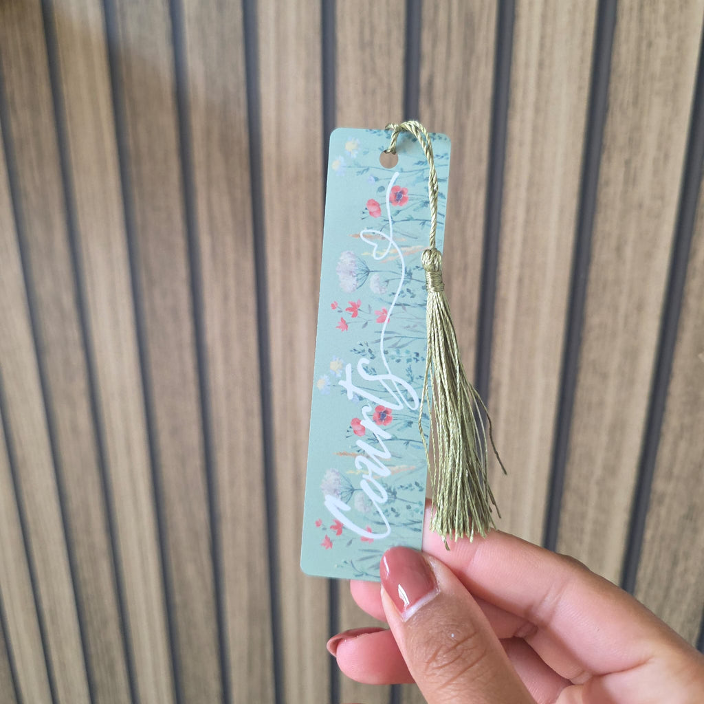 A hand with pink French-tipped nails holds a light blue rectangular bookmark featuring white cursive text and a floral pattern, with a sage green tassel attached at the top, set against a wooden slatted background.