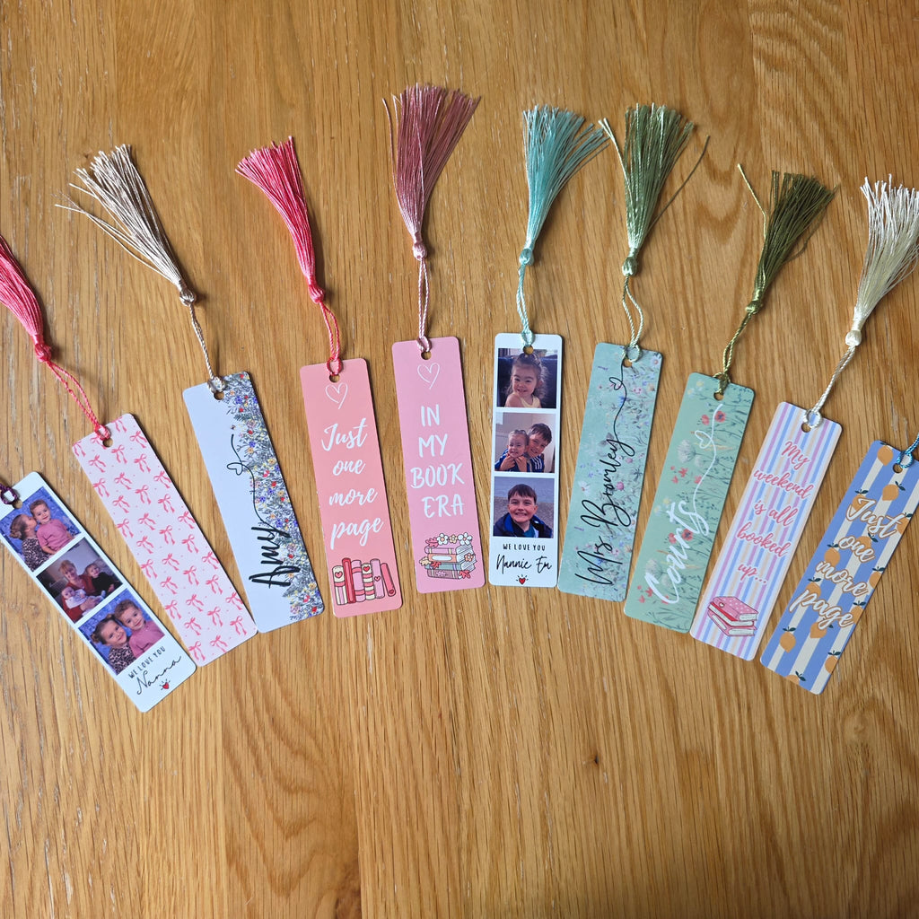 A row of personalized paper bookmarks with tassel tops laid out on a wooden tabletop. The bookmarks feature photos of a person, handwritten names, quotes like 'Just one more page' and 'IN MY BOOK ERA', and decorative patterns in pink, blue, green, and white.