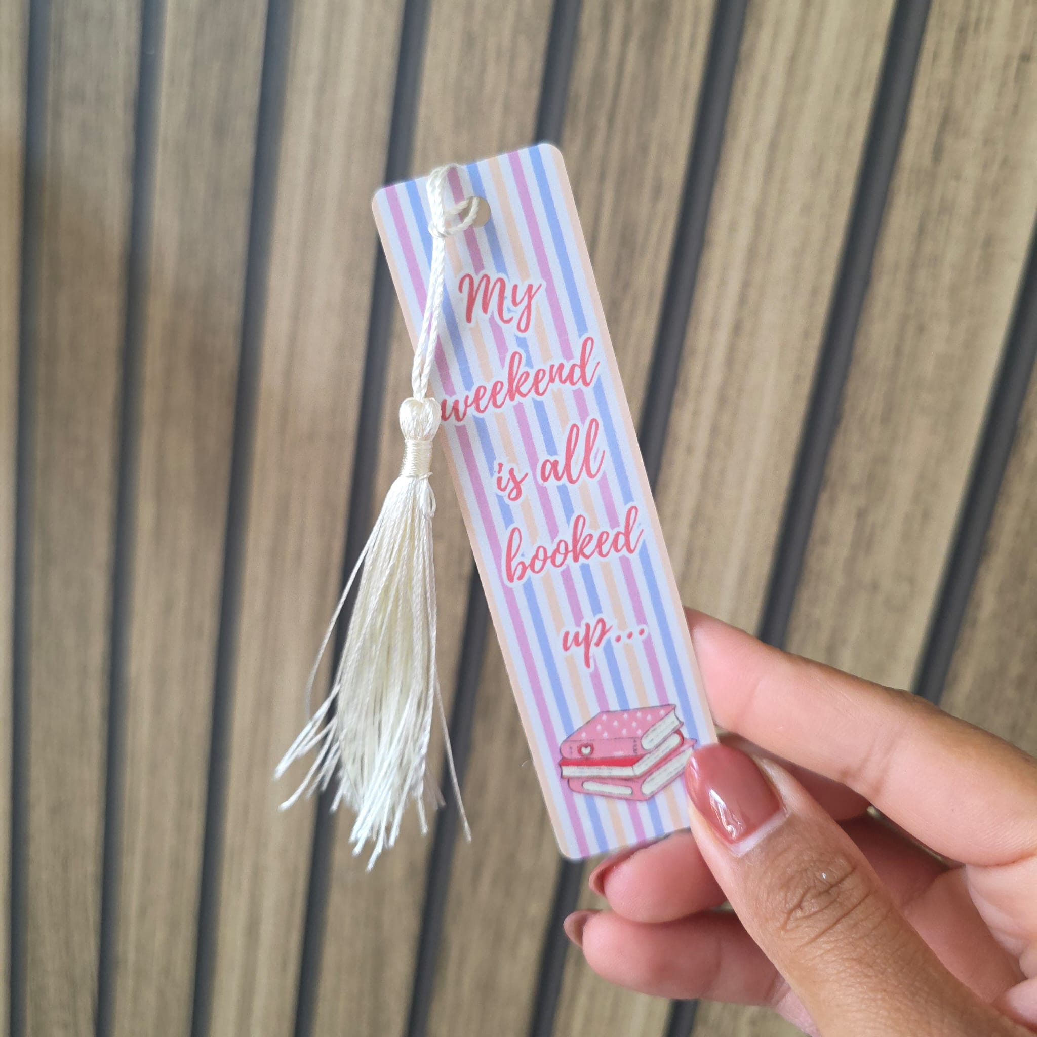 A hand holding a pastel striped paper bookmark with a white tassel. The bookmark features pink script text reading 'My weekend is all booked up.