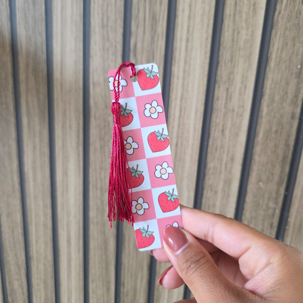 A hand holding a pink and white checkered bookmark with red tassels and strawberry and flower illustrations, set against a wooden slat background.