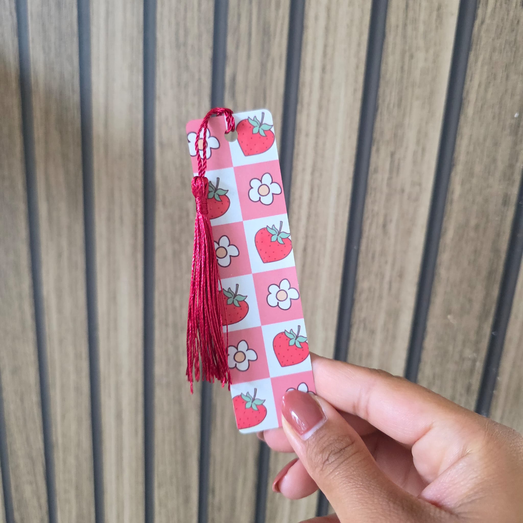 A hand holding a pink and white checkered bookmark with red tassels and strawberry and flower illustrations, set against a wooden slat background.
