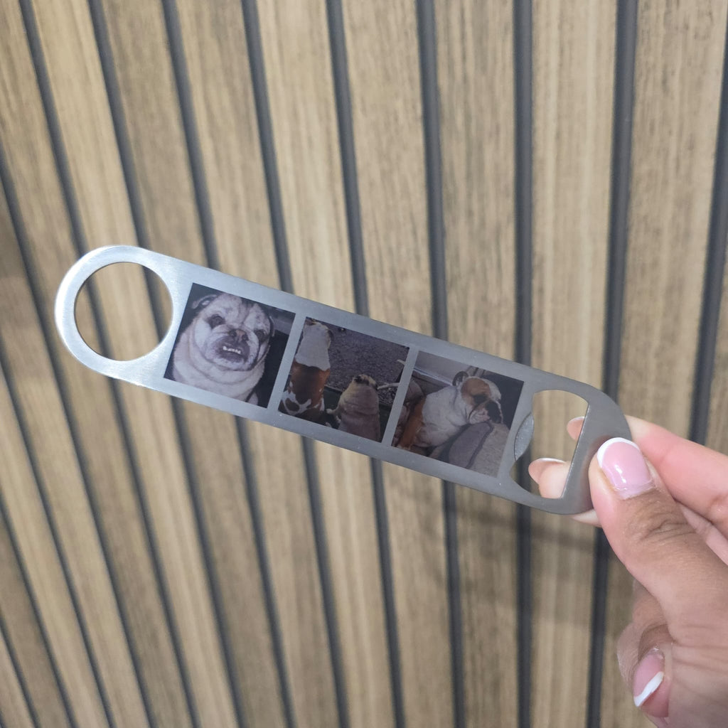 A hand holding a silver metal bottle opener engraved with a four-panel dog photo collage, set against a wooden slat background.