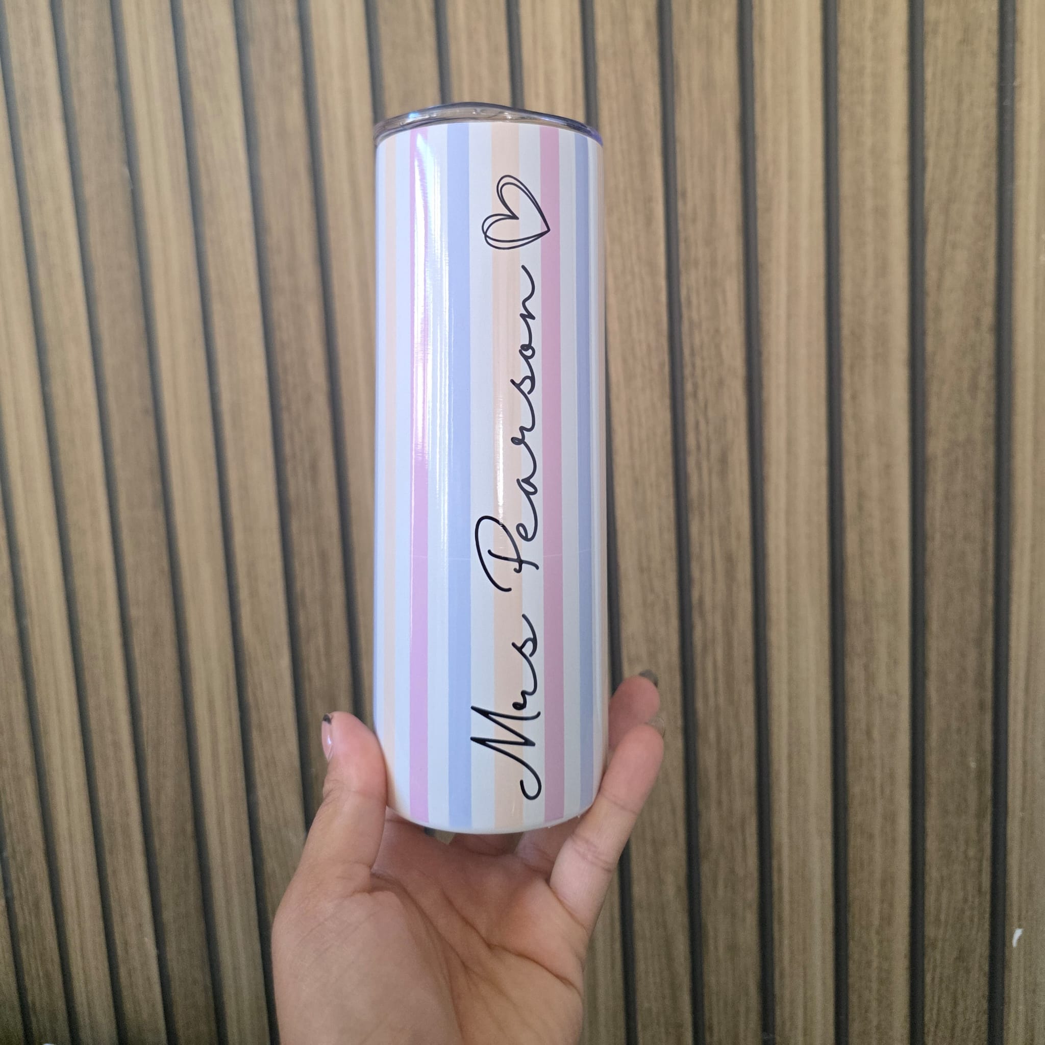 A hand holding a slim stainless steel tumbler with pastel vertical stripes in pink, blue, and white, featuring the text 'Miss Patterson' in black script with a heart above it, set against a wooden slat background.