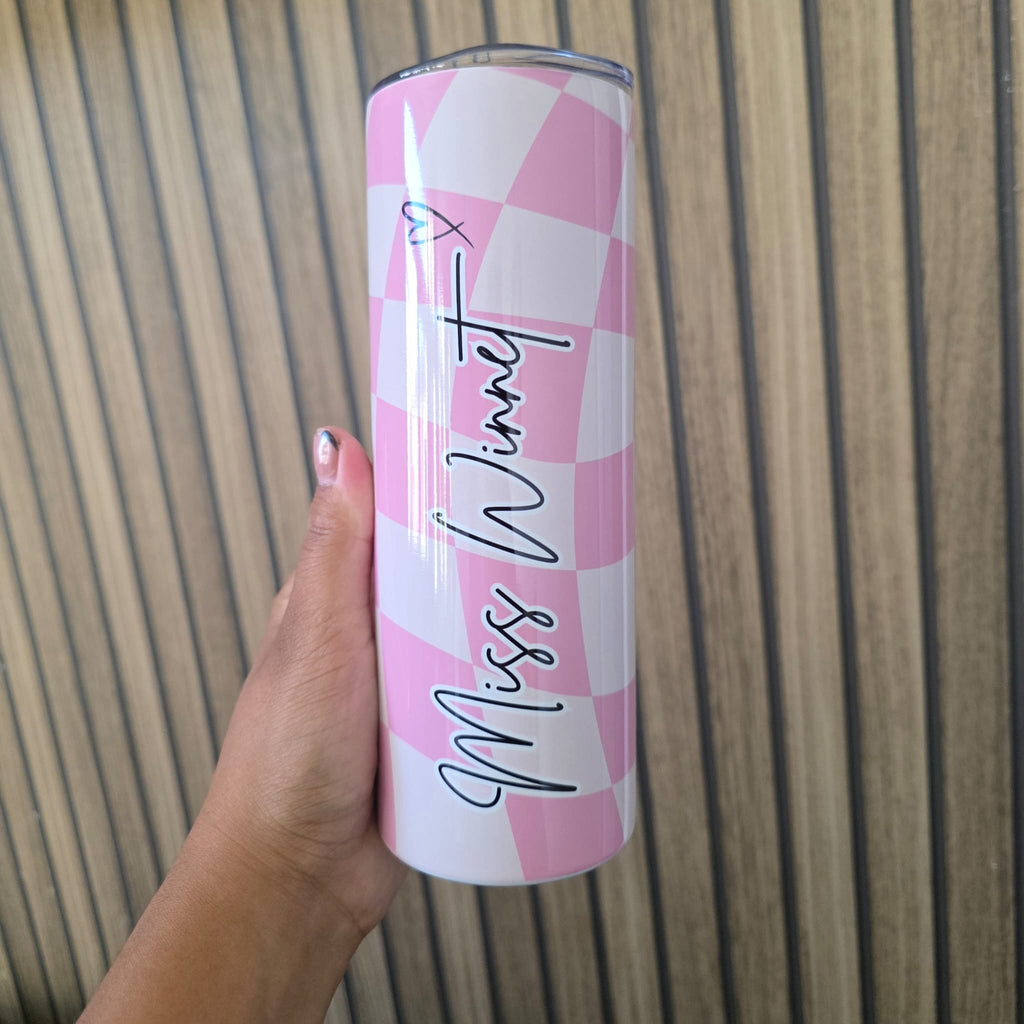 A hand holding a tall insulated tumbler with a pink and white checkerboard pattern and the text 'Missy Winet' written vertically in black script, set against a wood slat background.