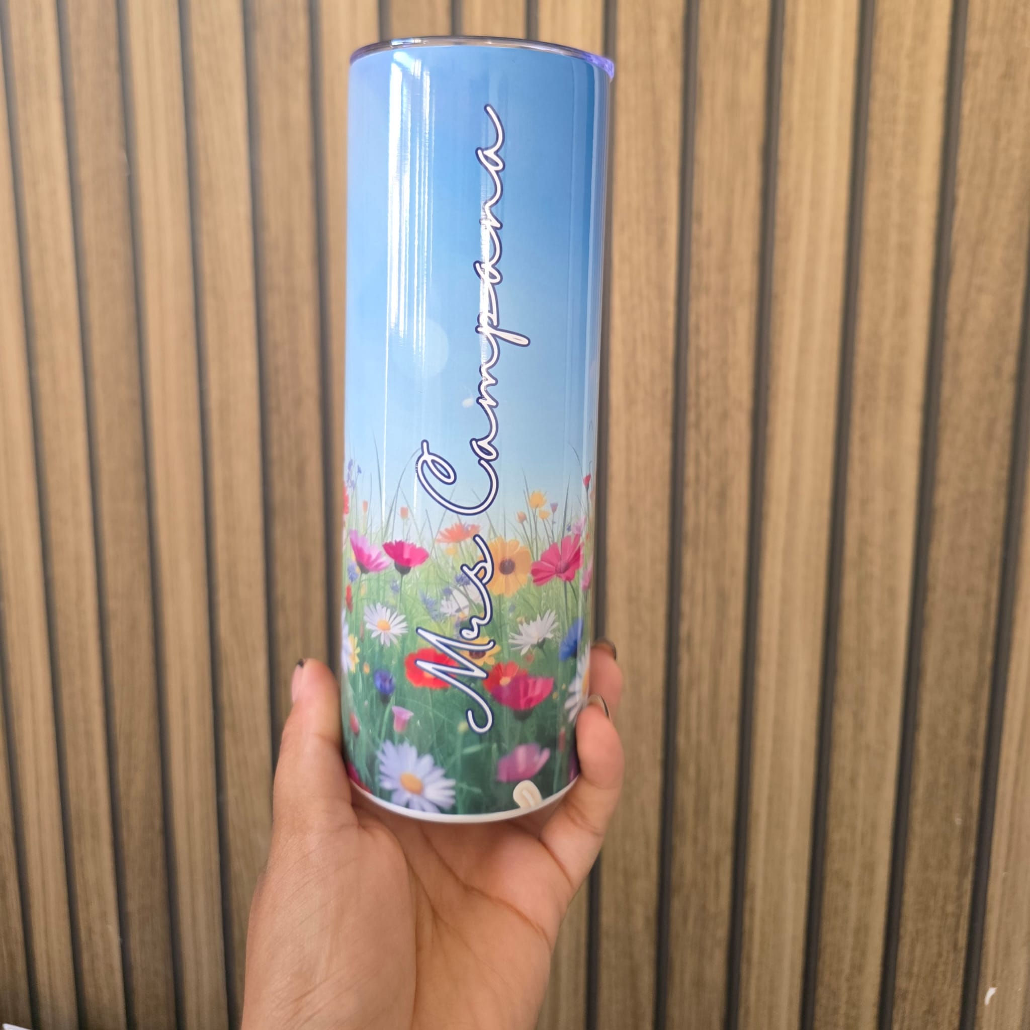 A hand holding a cylindrical metal tin with a blue sky and wildflower design, featuring the text 'Miss Campina' in white script, set against a wooden slat background.