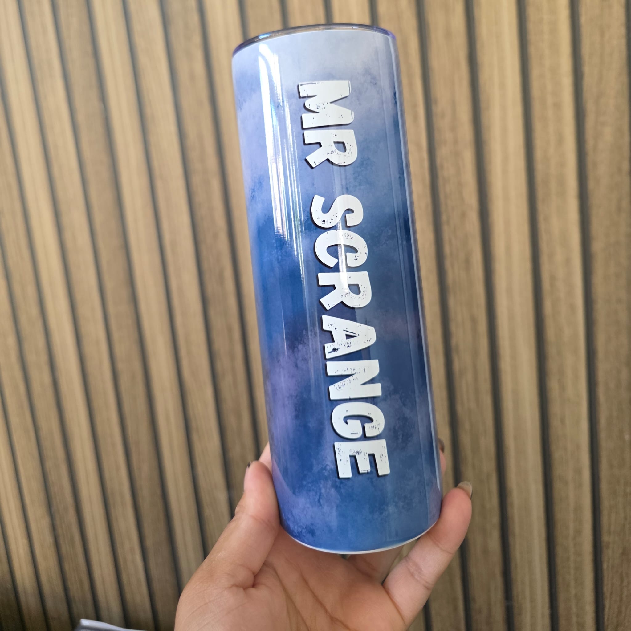 A hand holding a blue gradient insulated tumbler with 'MR SCRAMBLE' printed vertically in white text, set against a wooden slatted background.