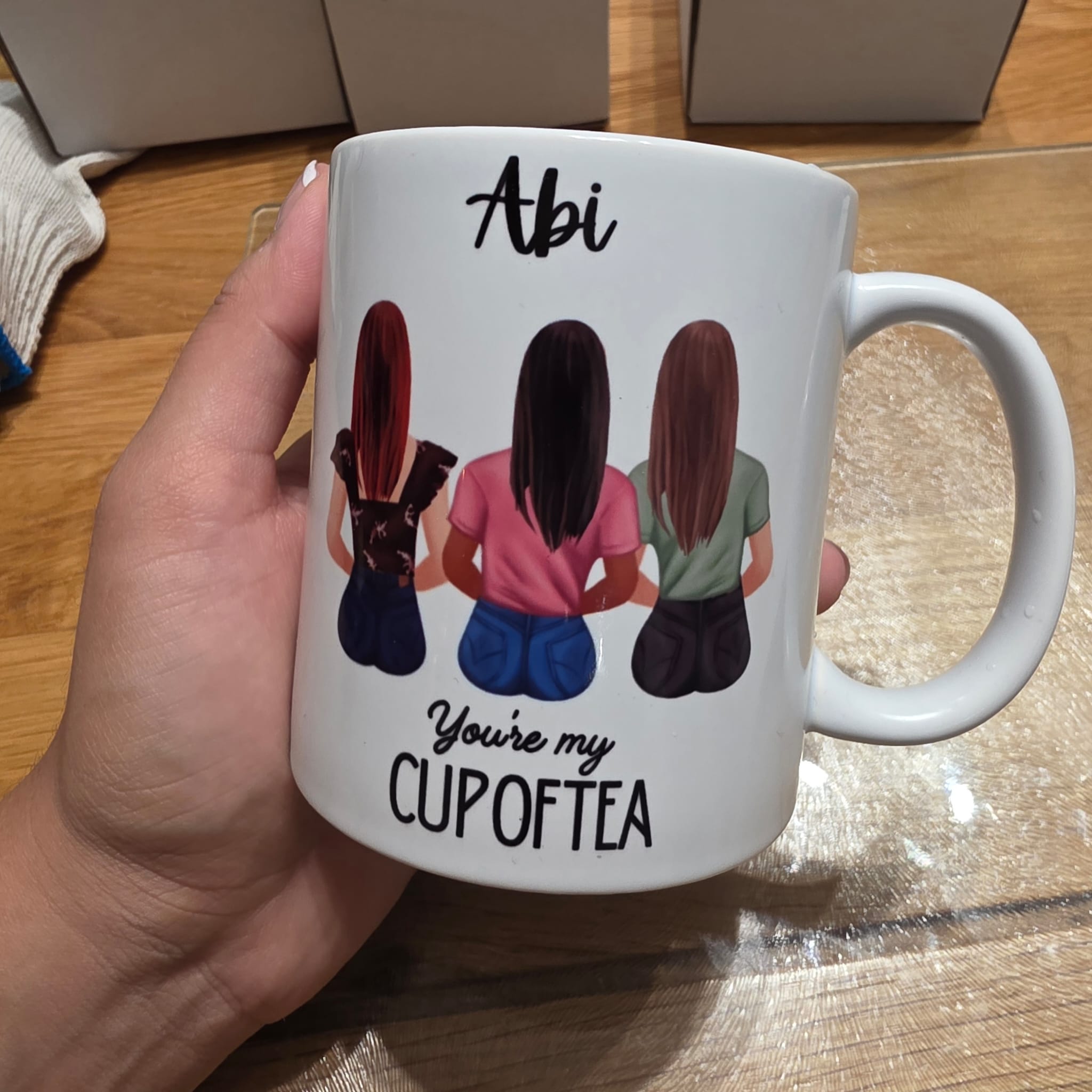 A hand holds a white ceramic mug with a printed illustration of three women from behind and the text 'Abi You're my CUP OF TEA'. The mug is shown on a wooden table.