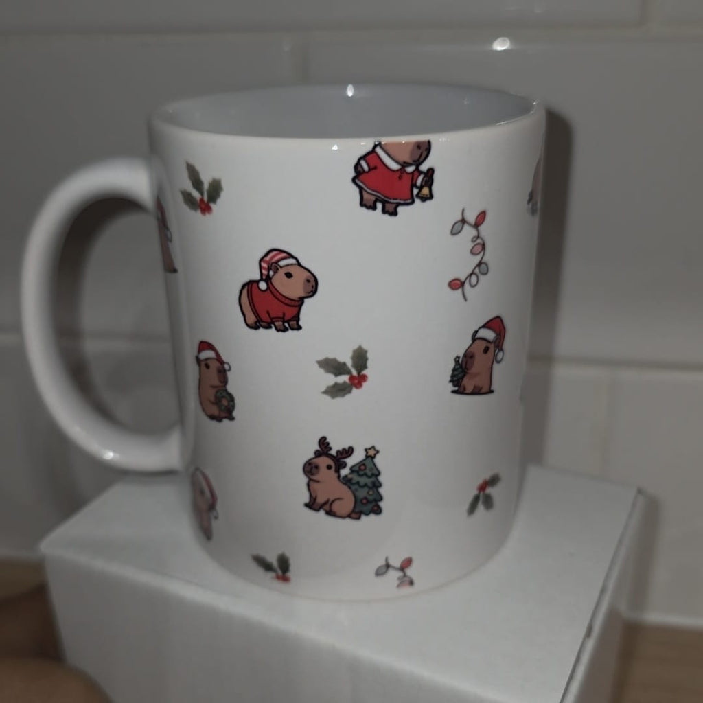 A white ceramic mug with a handle, decorated with cute cartoon capybara characters wearing Santa hats and holiday holly motifs, displayed on a white block against a tiled background.