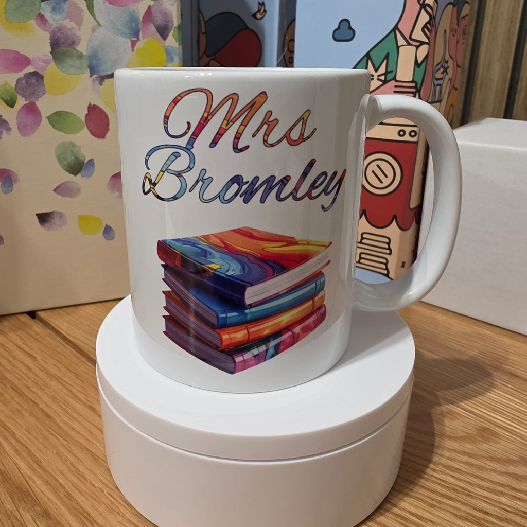A white ceramic mug with a colorful printed design featuring the text 'Mrs Bromley' in a rainbow gradient and an illustration of a stack of books, displayed on a white circular pedestal.