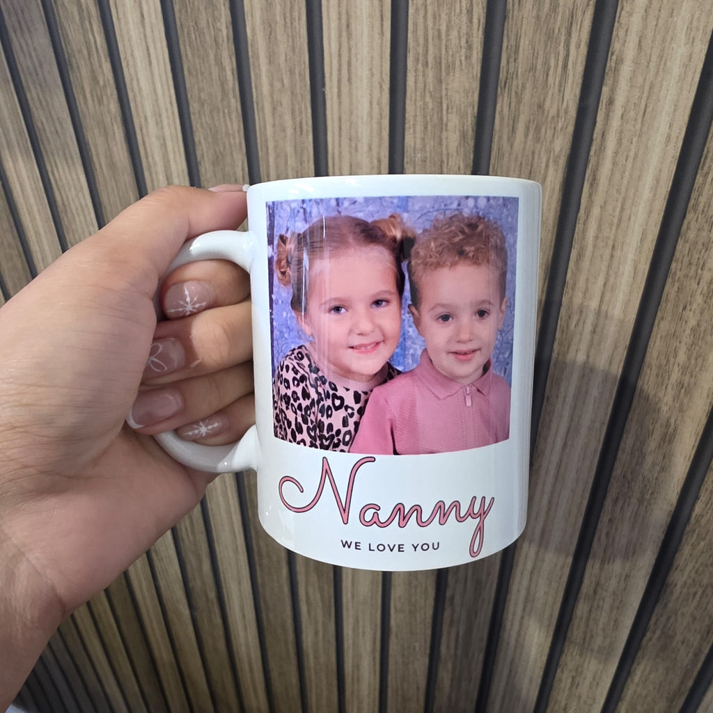 A hand holds a white ceramic mug with a custom photo of two children and the text 'Nanny WE LOVE YOU' against a wood-paneled background.