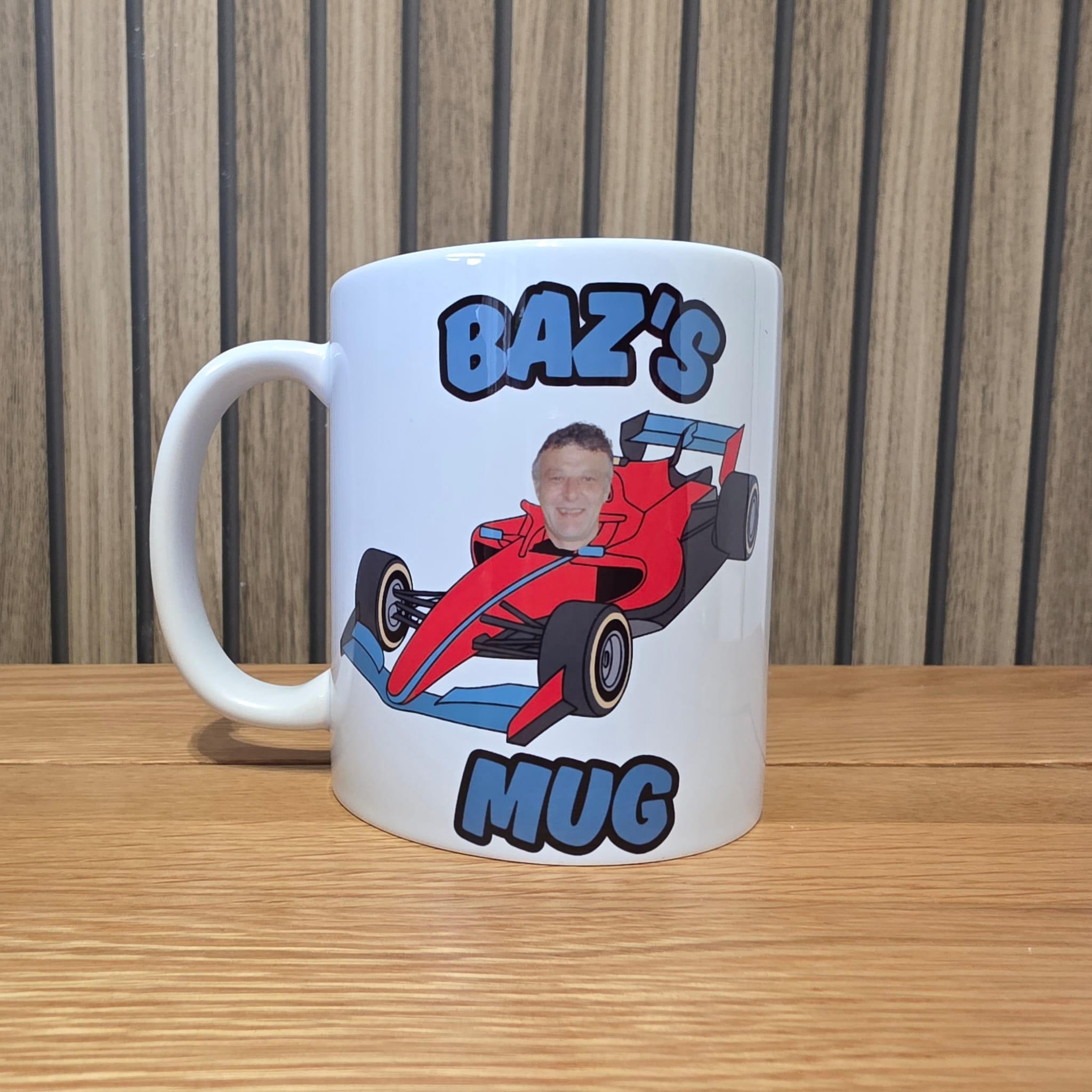 A white ceramic mug with a custom graphic print featuring a red race car and the text 'BAZ'S MUG' in blue and black, sitting on a wooden table against a wood-paneled background.
