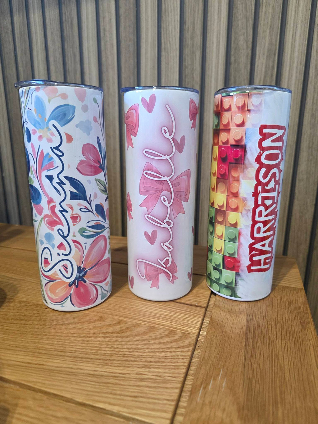 Three personalized tumblers with floral, name, and colorful pattern designs on a wooden surface.