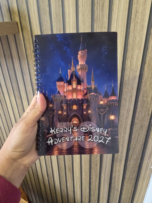 Notebook with a castle design and 'Kerry's Disney Adventure 2027' text held by a hand against a striped background.