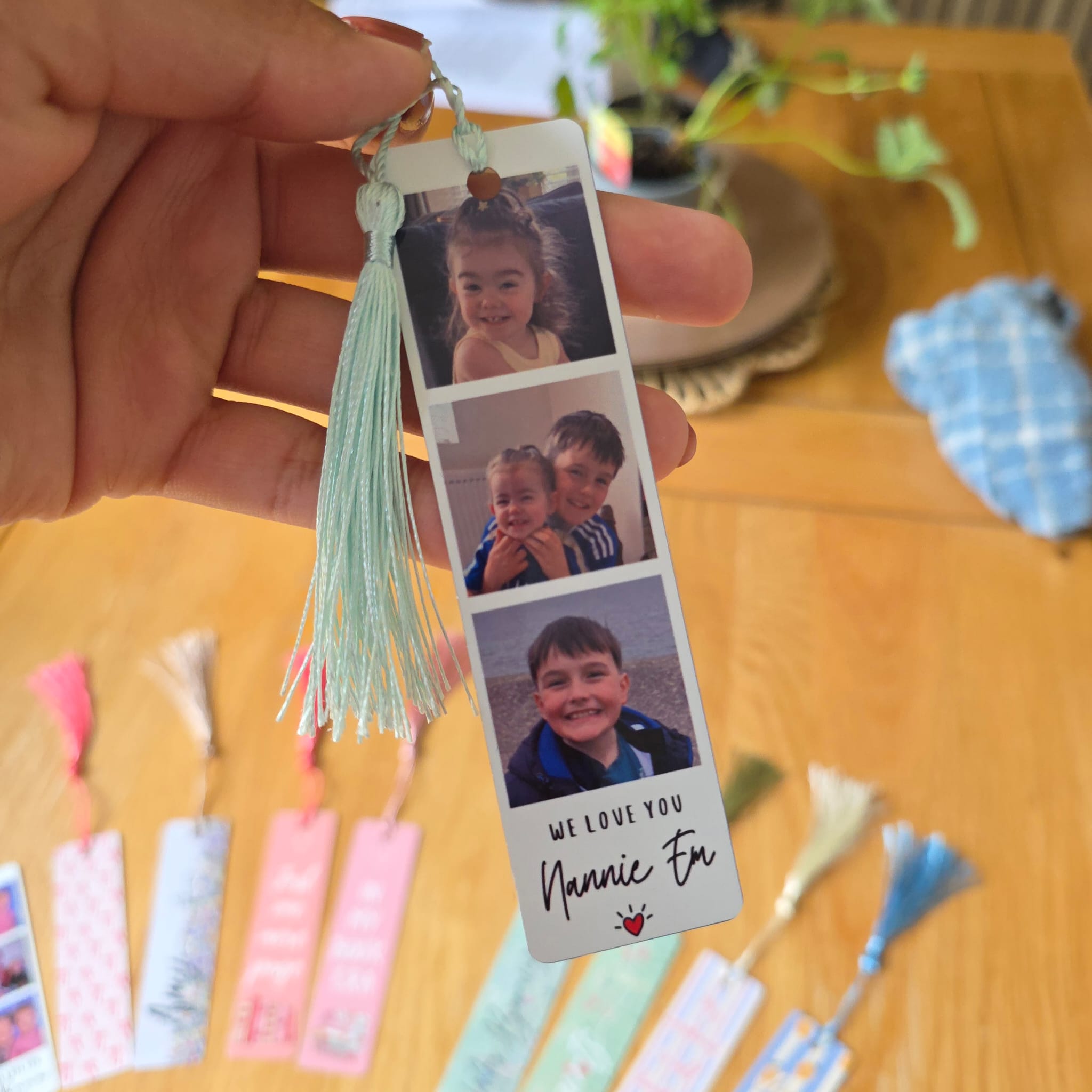 A hand holds a personalized photo bookmark featuring three printed child photos, a mint green tassel, and the text 'WE LOVE YOU Nannie Et' on a wooden table with other bookmarks blurred in the background.